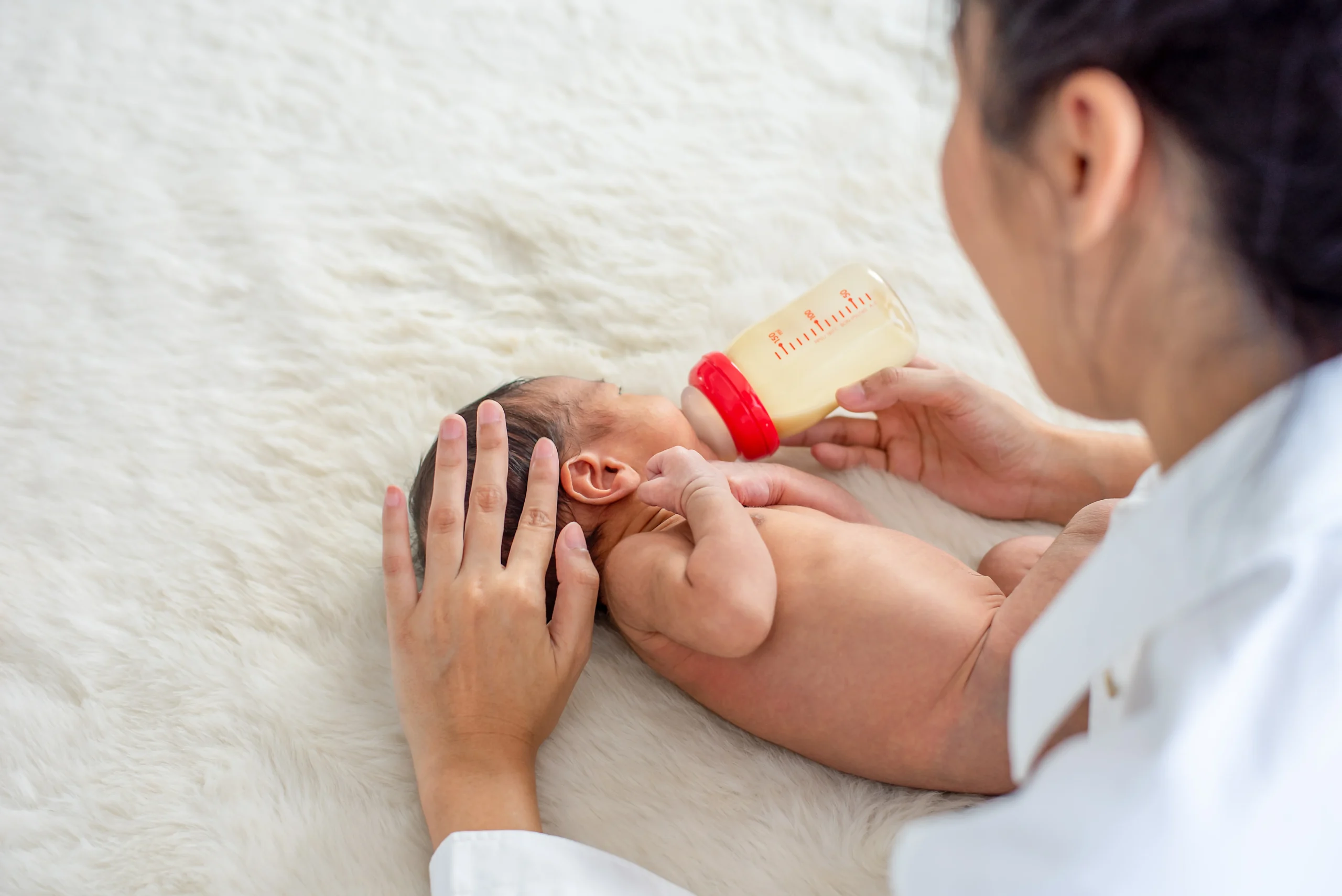 Asian mother give milk feeding with bottle to little newborn baby on white bed and take care - Neonatal Nutrition - Importance, Benefits & Myths - Kare One NICU Care